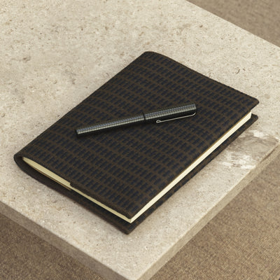 The Kelly Rutherford Limited Edition Notebook from August Sandgren, featuring Danish design and crafted with high-quality woven wool by Kjellerup Væveri. The notebook, shown in a dark pattern, includes Oeko-Tex certified leather and replaceable A5 paper inserts, reflecting Kelly Rutherford’s distinctive style in this Limited Edition Collection of handcrafted storage solutions.